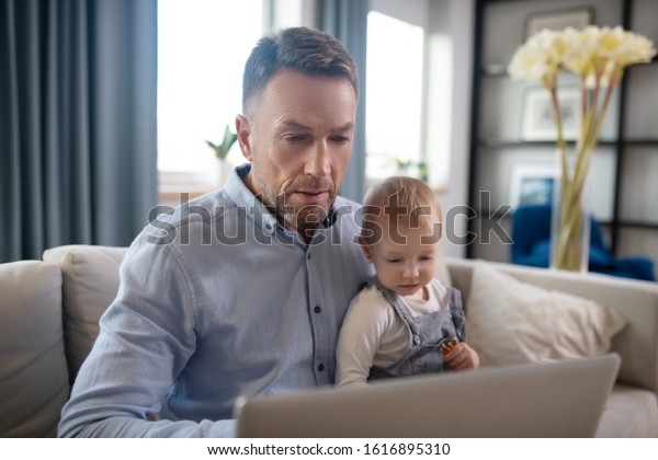 Busy father. Mature father holding his kid and\
working on his laptop