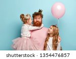 Busy father organizes holiday on International childrens day for two daughters, have home party, wear crowns and festive clothes. Little adorable girl holds air balloon, looks at dad and sister