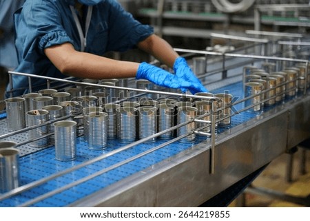 In a busy food processing facility, workers in blue clothing and gloves meticulously handle cans on a production line, ensuring quality and efficiency during daytime operations.