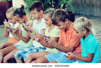 Busy glad children holding smartphones in hands and sitting together outdoors