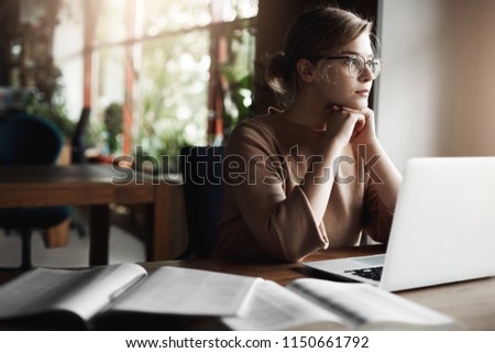 Busy and good-looking european female in glasses, leaning on hands, sitting in cafe while working remotely via laptop. Woman got carried away with thoughts in middle of studying, gazing through window
