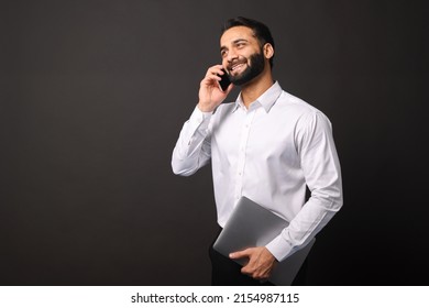 Busy Handsome Confident Bearded Indian Man In Formal Wear Talking On Smartphone Carrying Laptop Isolated On Black, Multiracial Male Entrepreneur Has Taking A Call On The Mobile Phone