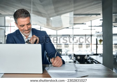 Busy happy middle aged smiling professional business man company executive ceo manager or lawyer wearing suit sitting at desk in modern office working on laptop computer writing notes, copy space.