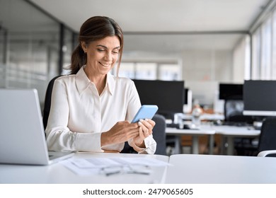 Busy happy professional middle aged business woman using mobile phone working in office. Smiling mature executive holding cell at desk. Happy female worker making payments on smartphone. Copy space. - Powered by Shutterstock