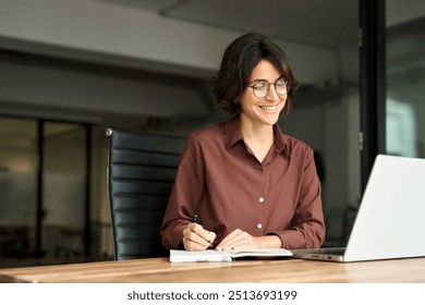 Busy happy young Hispanic business woman company owner ceo working in office using laptop writing notes. Female professional executive manager in her 30s using computer, elearning at workplace. - Powered by Shutterstock