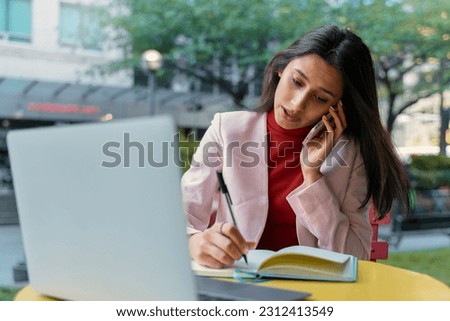 Busy Indian freelancer, young serious businesswoman talking on mobile phone, using laptop computer, taking notes, brainstorming, planning project working online at workplace. Multitasking concept 