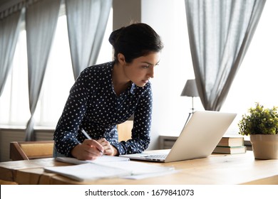 Busy Indian woman standing at table, writing notes in notebook with pen, focused female student using laptop, studying online at home, looking at screen, reading important information