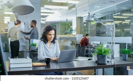Im Büro beschäftigt, schöne hispanische Frau, die an ihrem Schreibtisch auf einem Laptop arbeitet, im Hintergrund haben ihre Mitarbeiter Arbeitsgespräche. Stilvolles Büro mit hellen jungen Leuten.