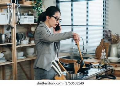 Busy Korean Female Worker Holding Spoon Is Frying Egg And Dealing With Work On Phone In The Morning. Asian Businesswoman Using Spatula And Pan Is Calling Her Boss And Cooking Breakfast For Family.