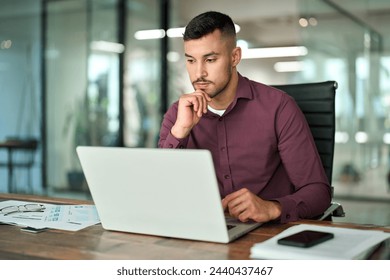 Busy Latin business man working on laptop sitting at office desk. Professional businessman company employee looking at computer thinking on online investment risks analyzing online financial data.