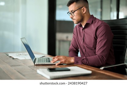 Busy Latin young business man executive working on laptop sitting at office desk. Professional serious businessman worker, male company employee manager looking at computer technology at workplace.