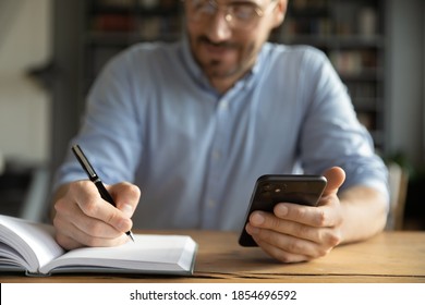 Busy male entrepreneur businessman working sitting at workplace desk holding smartphone writing schedule in diary with pen, close up image. Creates, planning to-do list in personal organizer concept