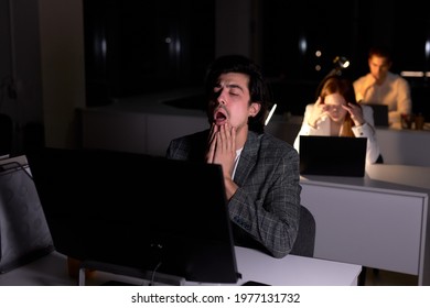 Busy Male Yawning While Working On Pc Computer, In Office At Night. Portrait Of Guy In Formal Wear Having Deadline, No Time To Sleep. Colleagues In The Background