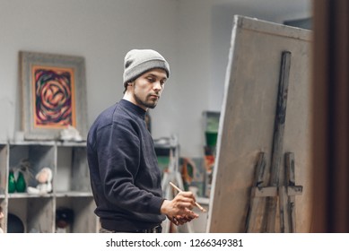 Busy man artist with a brush in his hand picks up an oil painting in a cozy studio. Talented young painter paints a painting on canvas. Painting Concept. Work as a hobby. - Powered by Shutterstock