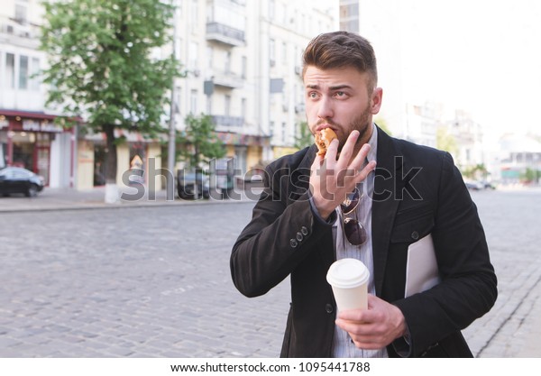 Busy man with a\
cup of coffee and a sandwich in his hands is in a hurry to work. A\
businessman is standing by the road, eating a sandwich and drinking\
coffee. Snack by fast\
food.
