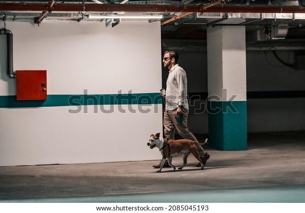 A busy man dressed smart casual\
is searching for his car in the underground garage with his dog.\
The man is holding coffee to go and passing by the\
wall.