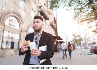 A Busy Man Eats And Drinks Coffee While Walking For Work. Business Man Has Breakfast With Fast Food. A Businessman Goes To Work On The Street And Eats A Sandwich