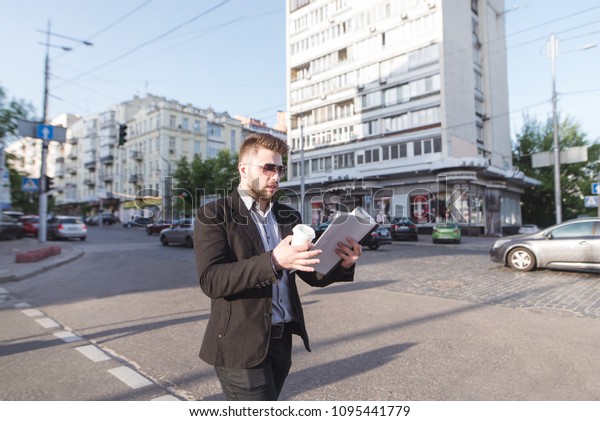 Busy man passes the road at the traffic light,
looks at the documents in his hands and drinks coffee from the cup.
Business man goes to work.