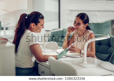 Busy master. Long-haired professional nail master talking to her curious client while taking care of her nails