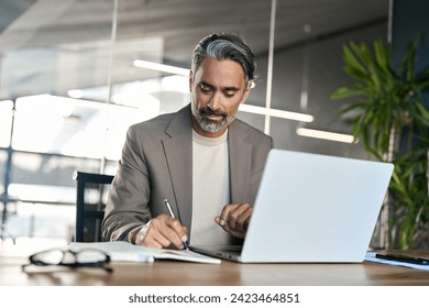 Busy mature business man, middle aged professional executive manager lawyer wearing suit writing notes watching online webinar analysing financial data using laptop computer working in office at desk. - Powered by Shutterstock