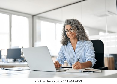 Busy mature business woman working in office using laptop writing. Smiling middle aged professional executive looking at computer, elearning, watching webinar having hybrid meeting sitting at desk.