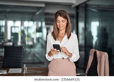 Busy mature elegant business woman model or mid age holding smartphone looking at cellphone standing in office. Middle aged female executive using cell phone digital tech device at work. - Powered by Shutterstock