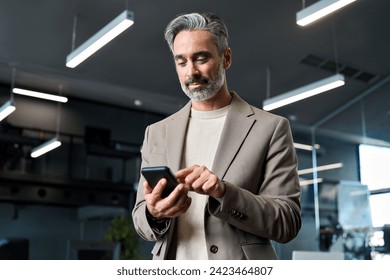 Busy mature older business man wearing suit using mobile cell phone at work. Middle aged businessman executive manager or entrepreneur holding smartphone looking at cellphone standing in office. - Powered by Shutterstock