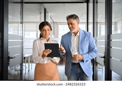 Busy mature professional business people, two happy business man and woman executives partners team working together walking in office using digital tablet talking on company financial project. - Powered by Shutterstock