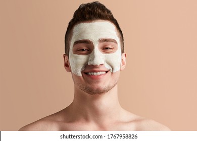 Busy Men Prefer Minimalist, Easy And Quick Facial Treatments. A Young Brown-haired Man With White Drying And Cracking Mask On His Unshaven Face Is Smiling And Looking Straight Into The Camera.