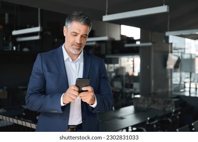 Busy mid aged business man ceo wearing blue suit standing in office using mobile cell phone. Mature businessman professional executive manager holding smartphone checking financial apps on cellphone. - Powered by Shutterstock