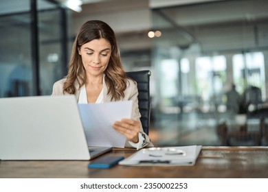 Busy mid aged business woman working in office with laptop reading document. Mature professional female manager lawyer attorney holding paper finance report sitting at desk in office. Copy space