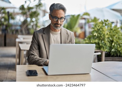 Busy middle aged business man executive ceo wearing suit and glasses sitting at outdoor table using laptop. Mature professional businessman manager hybrid working looking at computer outside office. - Powered by Shutterstock