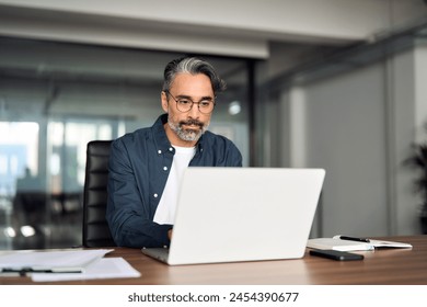 Busy middle aged business man executive wearing shirt and glasses sitting at desk using laptop. Serious mature professional businessman financial manager working online looking at computer in office. - Powered by Shutterstock