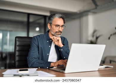 Busy middle aged business man executive wearing shirt and glasses sitting at desk using laptop. Mature serious professional businessman manager working looking at computer technology in office.