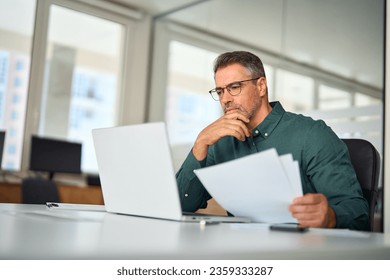 Busy middle aged executive, mature male hr manager holding documents using laptop looking at pc in office at desk, thinking over financial data report feeing doubt about market assets investment risk. - Powered by Shutterstock