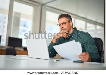 Busy middle aged executive, mature male hr manager holding documents using laptop looking at pc in office at desk, thinking over financial data report feeing doubt about market assets investment risk.