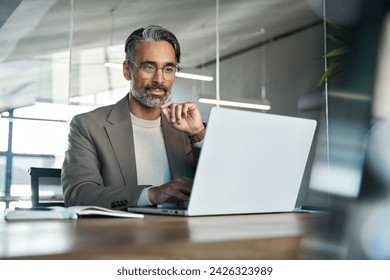 Busy middle aged Latin business man executive ceo wearing suit and glasses sitting at desk using laptop. Mature professional businessman manager working looking at computer technology in office. - Powered by Shutterstock