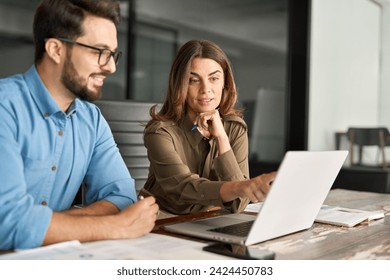 Busy middle aged Latin female manager explaining corporate software work talking to young male employee consulting client working on computer using laptop discussing business plan at office meeting. - Powered by Shutterstock