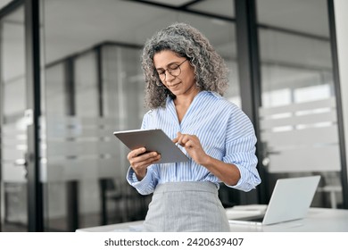 Busy middle aged professional business woman using tab computer in office. Mature senior businesswoman bank manager, older female corporate executive holding digital tablet standing at work. - Powered by Shutterstock