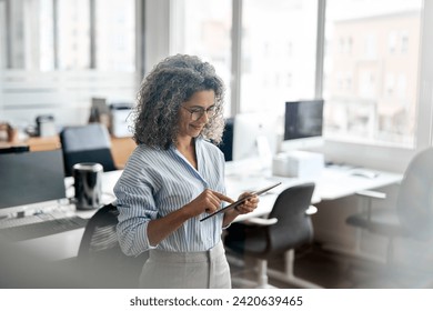 Busy middle aged professional business woman using tab computer in office. Mature lady manager, older female corporate executive holding tablet standing at work, authentic shot. View through glass - Powered by Shutterstock