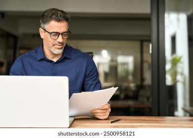 Busy middle aged professional business man using laptop computer reading document at work. Senior mature male manager executive working on laptop checking paper financial report in office. Copy space.