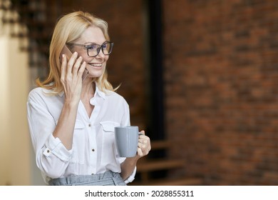 Busy middle aged woman with blonde hair and glasses in white shirt drinking coffee or tea while talking on her phone, standing at home - Powered by Shutterstock