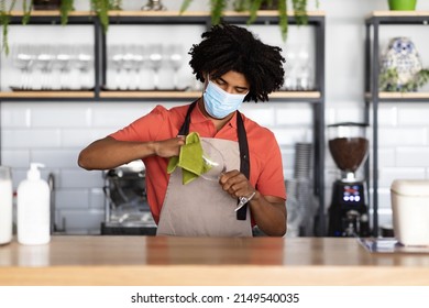 Busy Millennial Black Curly Male Waiter In Protective Mask And Apron Wipes Glass With Rag Behind Bar Counter, Work In Cafe Interior. Modern Business, Service, Startup, New Normal Due Covid-19 Outbreak