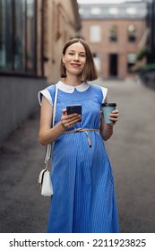 Busy Modern Pregnant Woman Walking Among Office Buildings Using A Smartphone. Woman In A Blue Vintage Dress And White Purse Using Phone On The Go