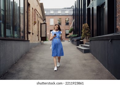 Busy Modern Pregnant Woman Walking Among Office Buildings Using A Smartphone. Woman In A Blue Vintage Dress And White Purse Using Phone On The Go