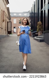 Busy Modern Pregnant Woman Walking Among Office Buildings Using A Smartphone. Woman In A Blue Vintage Dress And White Purse Using Phone On The Go