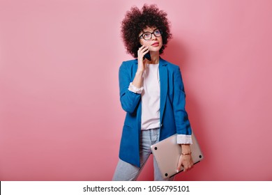 Busy modern pretty woman with ringlets wears glasses and blue jacket stands on isolated pink background, talking on smartphone and holds laptop, business woman 