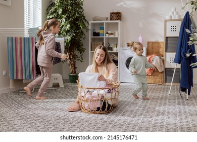 Busy mom sits on bathroom floor in laundry room and sorts colorful white clothes in wicker basket, washing machine in the background, girls running around, fun, joy, laughter, small children. - Shutterstock ID 2145176471