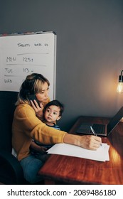 Busy Mom Writing Notes During A Business Phone Call. Working Mom Carrying Her Son On Her Lap While Sitting In Her Home Office. Mother Of One Making Business Plans While Working Remotely.