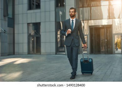 Busy morning. Full length of young and handsome bearded businessman in suit pulling his luggage and holding cup of coffee while walking outdoors - Powered by Shutterstock